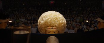 Movie still from “Isle of Dogs” (2018), directed by Wes Anderson – A person with a wig and a microphone in front of an audience; Extreme Close Up shot, Low angle