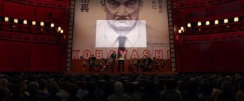 Movie still from “Isle of Dogs” (2018), directed by Wes Anderson – A group of people standing in front of a large screen; Extreme Wide shot, Low angle