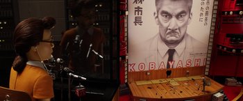 Movie still from “Isle of Dogs” (2018), directed by Wes Anderson – A man sitting in front of a poster of a man; Wide shot, High angle