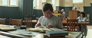 Movie still from “It” (2017), directed by Andy Muschietti – A young boy is reading a book at a table; Medium shot, Low angle
