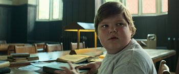 Movie still from “It” (2017), directed by Andy Muschietti – A young boy sitting in front of a table with a book; Medium shot, Low angle