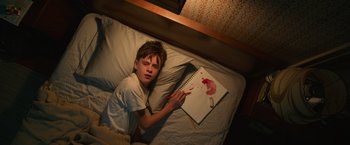 Movie still from “It” (2017), directed by Andy Muschietti – A boy laying in a bed with a book; Medium shot, Overhead angle