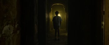 Movie still from “It” (2017), directed by Andy Muschietti – A person standing in a dark hallway with a light shining on them; Wide shot, Low angle