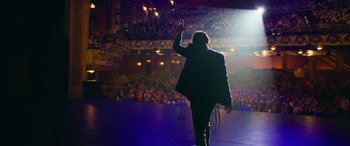 Movie still from “It Chapter Two” (2019), directed by Andy Muschietti – A man in a black jacket is on a stage waving; Wide shot, High angle