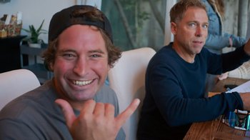 Movie still from “Jackass 4.5” (2022), directed by Jeff Tremaine – Two men sitting in a chair and one of the men is making a hand sign; Medium shot, Over the shoulder angle