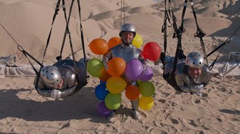 Movie still from “Jackass 4.5” (2022), directed by Jeff Tremaine – A man in a suit of armor and a helmet is holding a bunch of balloons; Wide shot, High angle