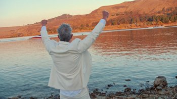 Movie still from “Jackass 4.5” (2022), directed by Jeff Tremaine – An older man standing on the shore of a body of water with his arms in the air; Medium shot, Low angle
