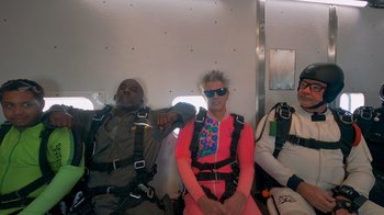 Movie still from “Jackass 4.5” (2022), directed by Jeff Tremaine – A group of people sitting on a plane wearing parachuting gear; Wide shot, High angle
