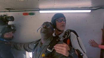 Movie still from “Jackass 4.5” (2022), directed by Jeff Tremaine – A man in a helmet is holding on to another man in a parachute; Medium shot, Overhead angle