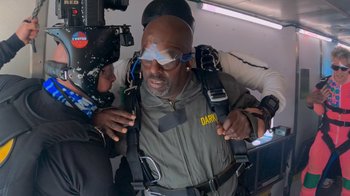 Movie still from “Jackass 4.5” (2022), directed by Jeff Tremaine – An older man in a parachute is being assisted by a cameraman; Medium shot, Over the shoulder angle