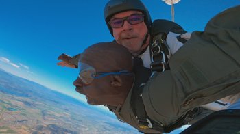 Movie still from “Jackass 4.5” (2022), directed by Jeff Tremaine – A man in a helmet is holding onto another man in a parachute; Medium shot, Overhead angle