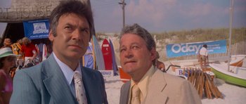 Movie still from “Jaws 2” (1978), directed by Jeannot Szwarc – Two men in suits standing in front of a row of surfboards on the beach; Close Up shot, Low angle