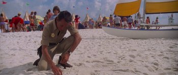 Movie still from “Jaws 2” (1978), directed by Jeannot Szwarc – A man kneeling on the beach with a surfboard in his hand; Wide shot, High angle