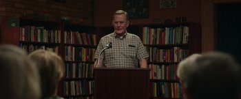 Movie still from “Jerry and Marge Go Large” (2022), directed by David Frankel – An older man standing at a podium in front of books; Medium shot, Low angle