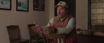 Movie still from “Jerry and Marge Go Large” (2022), directed by David Frankel – A man sitting in a chair with his hands outstreched; Medium shot, Over the shoulder angle