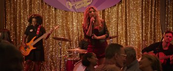 Movie still from “Jerry and Marge Go Large” (2022), directed by David Frankel – A woman singing into a microphone in front of a group of people; Wide shot, Over the shoulder angle