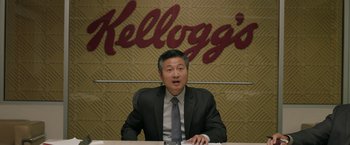Movie still from “Jerry and Marge Go Large” (2022), directed by David Frankel – A man sitting at a table in front of a kellogg's sign; Medium shot, Low angle