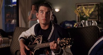 Movie still from “Jerry Maguire” (1996), directed by Cameron Crowe – A young man playing an acoustic guitar in a living room; Close Up shot, Over the shoulder angle