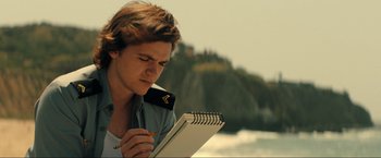 Movie still from “Jesus Revolution” (2023), directed by Brent McCorkle – A young man writing on a notepad near the ocean; Close Up shot, Low angle
