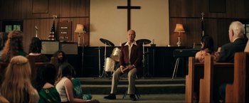 Movie still from “Jesus Revolution” (2023), directed by Brent McCorkle – A man sitting on a chair in front of a cross; Wide shot, Over the shoulder angle