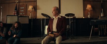 Movie still from “Jesus Revolution” (2023), directed by Brent McCorkle – An older man sitting in front of drums holding a book; Medium shot, Over the shoulder angle
