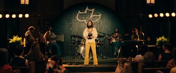 Movie still from “Jesus Revolution” (2023), directed by Brent McCorkle – A bearded man in a white shirt is on a stage; Wide shot, Low angle