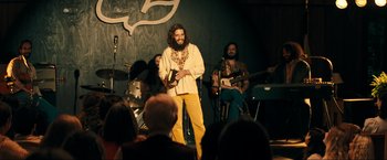 Movie still from “Jesus Revolution” (2023), directed by Brent McCorkle – A bearded man in a white shirt is on stage with a band; Medium shot, Over the shoulder angle