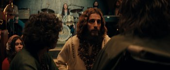 Movie still from “Jesus Revolution” (2023), directed by Brent McCorkle – A man with long hair and a beard is sitting in front of a group of people; Medium shot, Over the shoulder angle