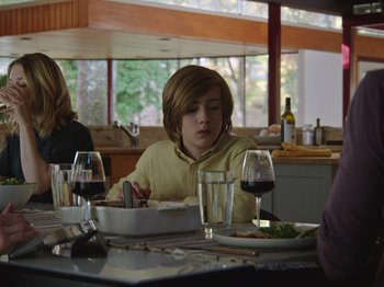 Movie still from “John and the Hole” (2021), directed by Pascual Sisto – A young boy sitting at a table with a plate of food; Medium shot, Over the shoulder angle