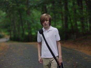 Movie still from “John and the Hole” (2021), directed by Pascual Sisto – A young man walking down a street holding a bag; Medium shot, High angle