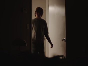 Movie still from “John and the Hole” (2021), directed by Pascual Sisto – A young boy standing in a room in front of a door; Medium shot, Over the shoulder angle