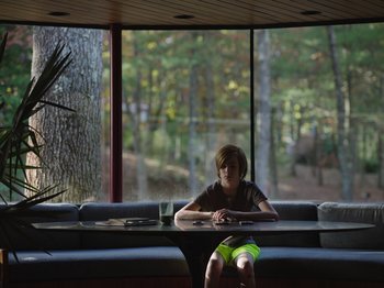 Movie still from “John and the Hole” (2021), directed by Pascual Sisto – A boy sitting at a table in front of a large window; Wide shot, Low angle