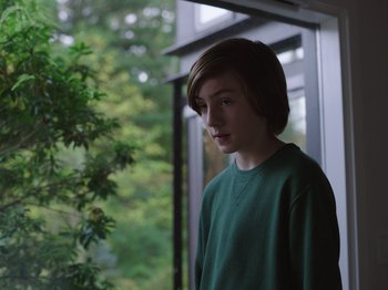 Movie still from “John and the Hole” (2021), directed by Pascual Sisto – A young boy standing in front of a window; Medium shot, Low angle