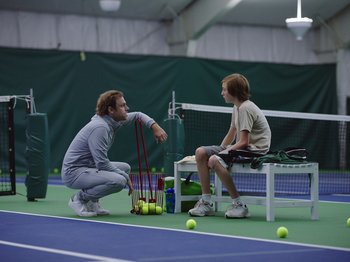 Movie still from “John and the Hole” (2021), directed by Pascual Sisto – Two men sitting on a tennis court with rackets and balls; Wide shot, Over the shoulder angle