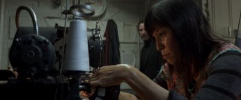 Movie still from “John Wick: Chapter 2” (2017), directed by Chad Stahelski – A woman working on a sewing machine in a room; Medium shot, Low angle