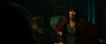 Movie still from “John Wick: Chapter 3 - Parabellum” (2019), directed by Chad Stahelski – A woman sitting at a wooden table in front of a painting; Medium shot, Over the shoulder angle