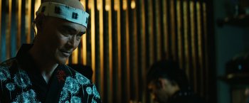 Movie still from “John Wick: Chapter 3 - Parabellum” (2019), directed by Chad Stahelski – A man wearing a paper hat standing next to another man in front of a wooden wall; Close Up shot, Low angle