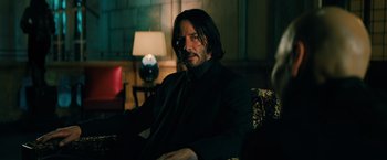 Movie still from “John Wick: Chapter 3 - Parabellum” (2019), directed by Chad Stahelski – A man sitting in a chair wearing a suit and tie; Medium shot, Over the shoulder angle