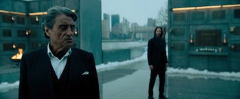 Movie still from “John Wick: Chapter 4” (2023), directed by Chad Stahelski – Two men standing on a sidewalk near a body of water; Medium shot, Low angle
