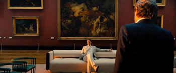 Movie still from “John Wick: Chapter 4” (2023), directed by Chad Stahelski – A man sitting on top of a white couch in front of a painting; Wide shot, Low angle