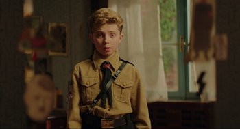 Movie still from “Jojo Rabbit” (2019), directed by Taika Waititi – A young boy in a uniform stands in front of a window; Medium shot, Over the shoulder angle