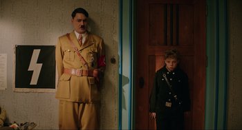 Movie still from “Jojo Rabbit” (2019), directed by Taika Waititi – A man and a boy dressed in military uniforms standing next to each other in a room; Medium shot, Over the shoulder angle