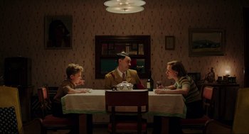 Movie still from “Jojo Rabbit” (2019), directed by Taika Waititi – A man sitting at a dinner table with two children; Wide shot, High angle