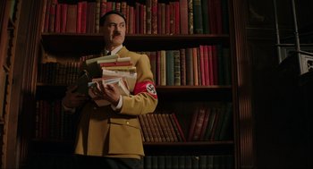 Movie still from “Jojo Rabbit” (2019), directed by Taika Waititi – A man in a suit holding a stack of books; Medium shot, Low angle