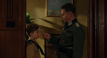 Movie still from “Jojo Rabbit” (2019), directed by Taika Waititi – A man in a military uniform is holding an object up to a young boy's face; Medium shot, Over the shoulder angle