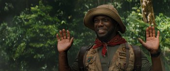 Movie still from “Jumanji: The Next Level” (2019), directed by Jake Kasdan – A man wearing a hat and a vest waving; Close Up shot, Over the shoulder angle