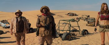 Movie still from “Jumanji: The Next Level” (2019), directed by Jake Kasdan – A man standing on top of a sandy beach next to a cart; Wide shot, High angle