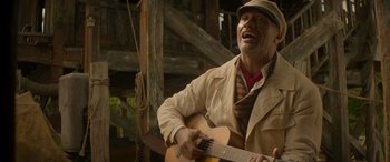 Movie still from “Jungle Cruise” (2021), directed by Jaume Collet-Serra – A man in a hat and a jacket is playing a guitar; Medium shot, Low angle