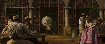 Movie still from “Jungle Cruise” (2021), directed by Jaume Collet-Serra – A person with an umbrella walking in front of a building; Wide shot, Over the shoulder angle