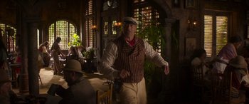 Movie still from “Jungle Cruise” (2021), directed by Jaume Collet-Serra – A man in a hat and vest standing next to a table; Wide shot, Over the shoulder angle